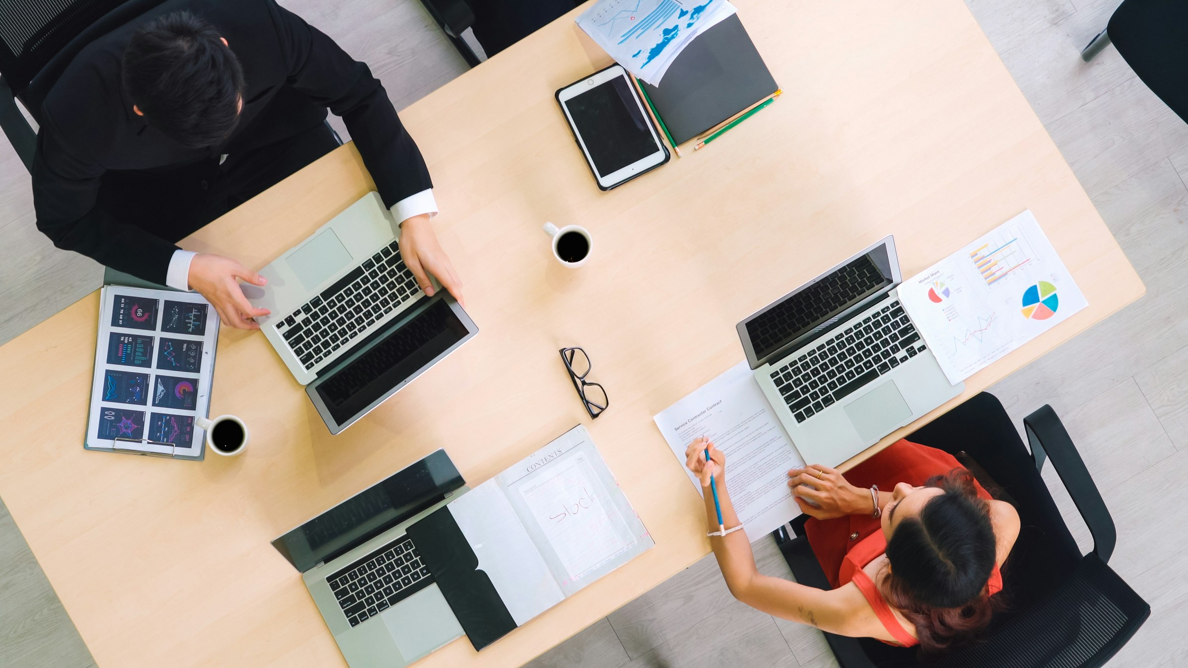 An overhead view of two professionals sitting at a light wood table, working on laptops surrounded by printed data dashboards, charts, and coffee mugs. One person is reviewing a document with a blue pencil, while the other manages a laptop next to a tablet and organized folders.